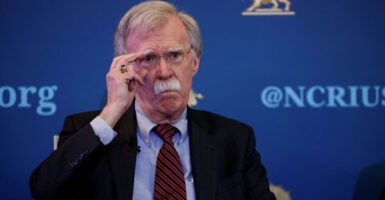 John Bolton in a blue suit with red-and-blue striped tie, adjusts his glasses.