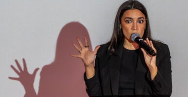 Alexandria Ocasio-Cortez in a black outfit holds a hand up, as her shadow is reflected on white wall behind her.