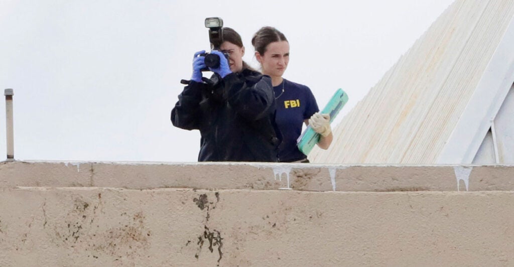 An FBI agent points a camera from the roof of a building, as another agent with documents in her hand stands behind her.
