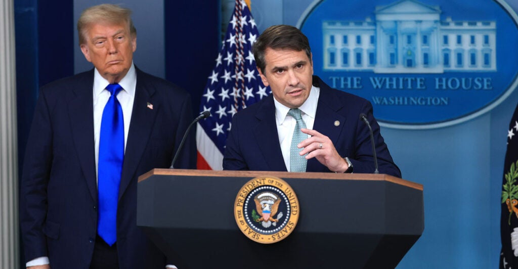 Deputy AG Todd Blanche speaks at the podium with President Donald Trump standing beside him.