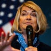Senator Cynthia Lummis of Wyoming speaks at a political event in front of an American flag back drop, holding a microphone.
