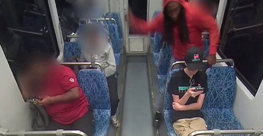 A screenshot of surveillance video showing the moment a man in a red shirt on a rail car begins to stab a young woman in a baseball cap, who is sitting looking at her cellphone.