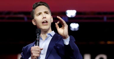 Sen. Josh Hawley in blue jacket and light-blue open shirt gestures as he speaks into a microphone.