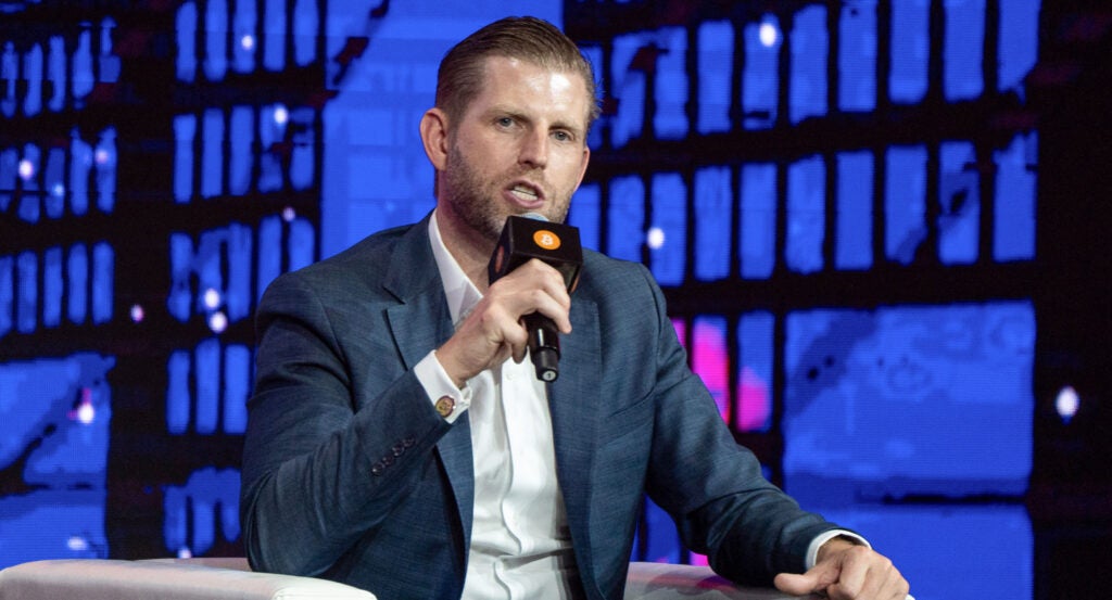 Eric Trump holds a microphone sitting on a white sofa and looking straight to the camera.