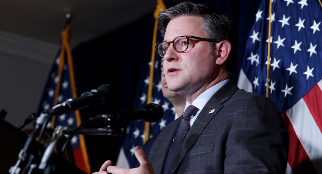 Mike Johnson looks out as he speaks into a bank of microphones, American flags behind him.
