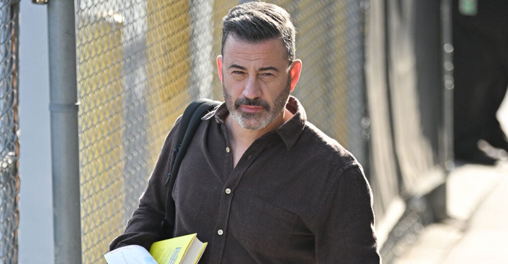 Jimmy Kimmel in a brown shit walking down a street, looking into the camera.