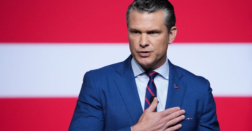 Pete Hegseth in a blue suit puts his hand over his heart.