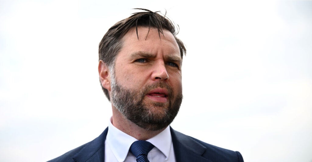 A close up photo shows JD Vance speaking, with a white sky behind him.