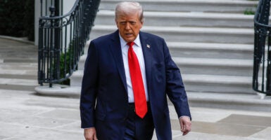 President Trump in a black suit and red tie walks in front of a staircase.