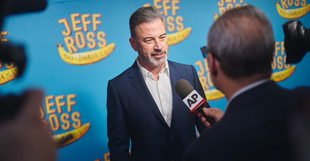 Jimmy Kimmel speaks to a reporter on a red carpet with blue background.