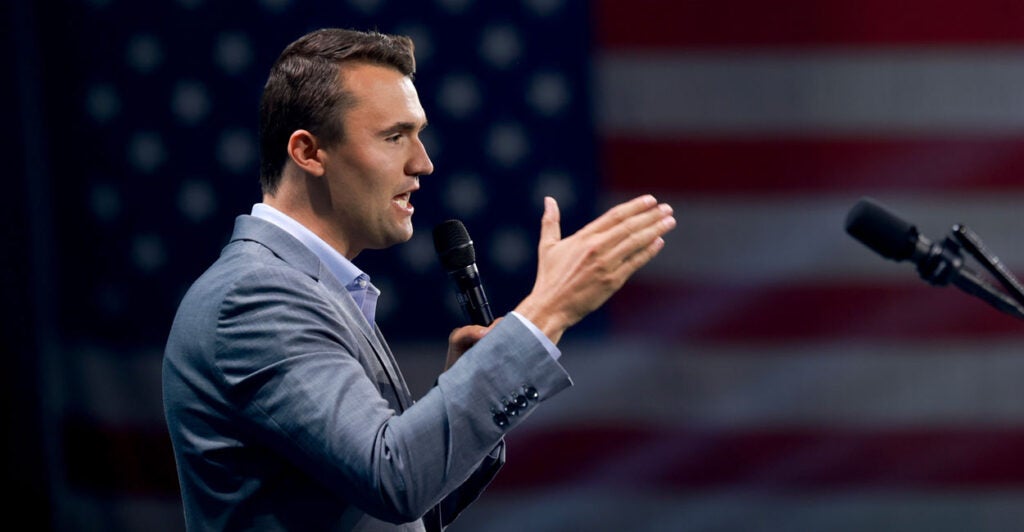 Charlie Kirk talking to an audience emphasizing with his hands with a large American flag in the background