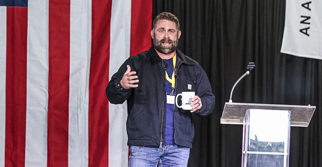 Nick Freitas in a pullover on stage talking to an audience in a warehouse with a large American flag in back