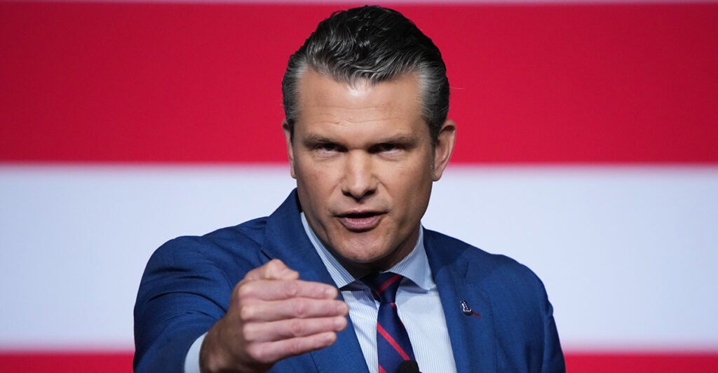 Pete Hegseth gestures toward the camera, with a large American flag as his backdrop.