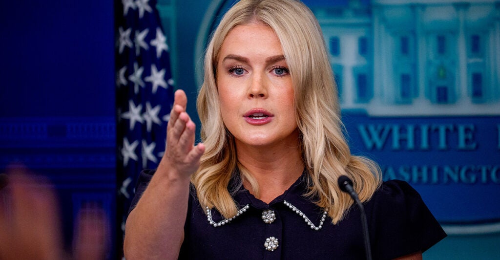 Karoline Leavitt in dark blue suit gestures with her right hand from White House podium.