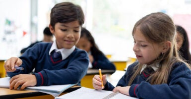 An eight-year-old boy and girl in blue sweater uniforms look over their notes in a private school classroom
