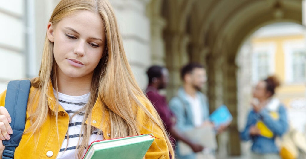A young worried-looking female student carrying a backpack and books while walking outside on campus with a group of students speaking to each other in the background