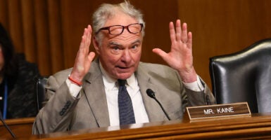 Senator Tim Kaine in a tan suit speaking at a Senate hearing, emphasizing with his hands raised and his glasses resting on his forehead
