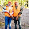 Father and son in orange hunting vests with hunting rifles in rural wooded area.