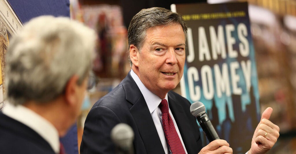 James Comey sits and holds a microphone with his right hand as he looks to his left.