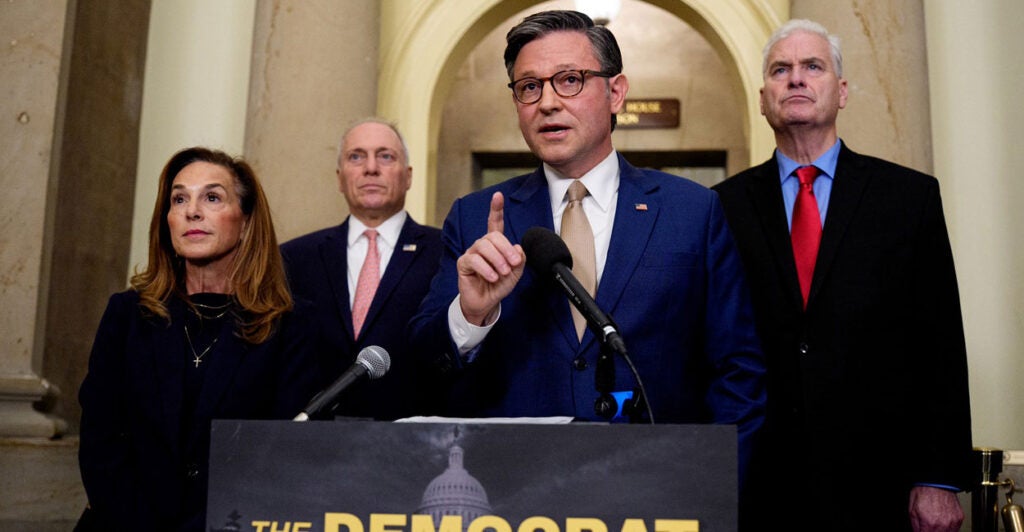 Mike Johnson stands at a podium and raises his right index finger as he speaks.