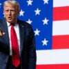 Donald Trump stands in front of an American flag as he points with his right hand.