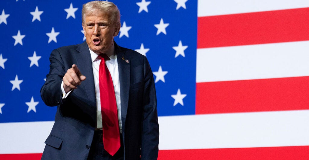 Donald Trump stands in front of an American flag as he points with his right hand.