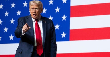Donald Trump stands in front of an American flag as he points with his right hand.
