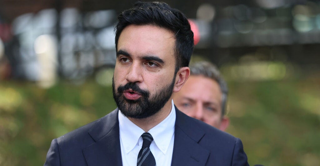 Zohran Mamdani stands outside at a press conference and looks to his right while answering questions.