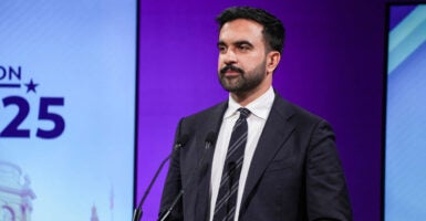 Zohran Mamdani stands behind a podium with a microphone in front of him and looks to his right.