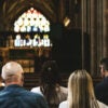A view of a stained-glass behind the podium at a church, with parishioners in the pews, as seen from behind.