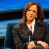 Kamala Harris sits on a chair with her hands folded together and a microphone in her right hand as she listens to a question.