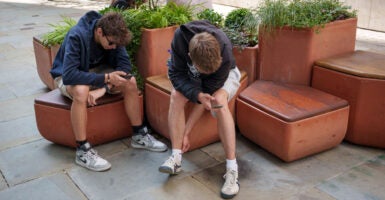 Two teen boys in black sweatshirts sit on brown chairs, oblivious to each other while staring at their smartphones.