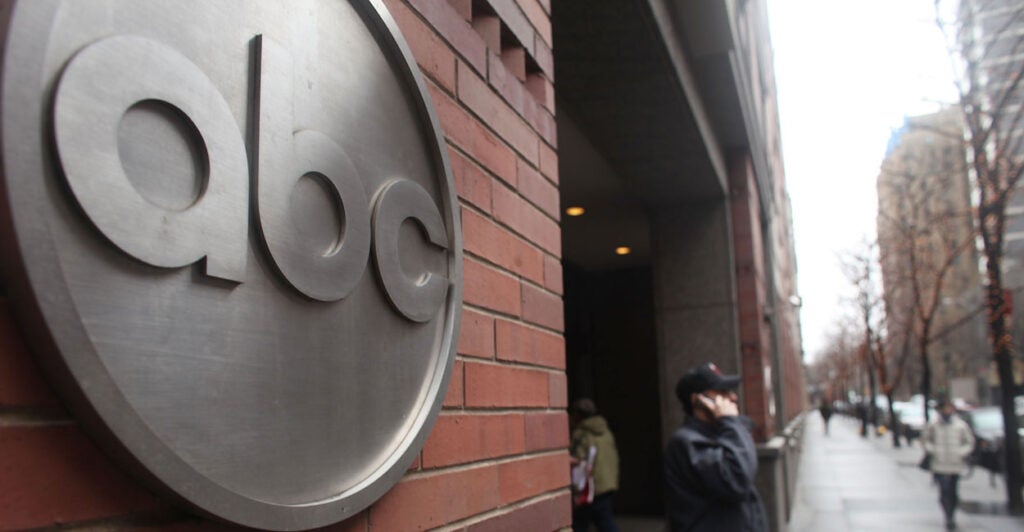 The ABC logo in silver on a brick building, as man stands with cellphone on sidewalk.