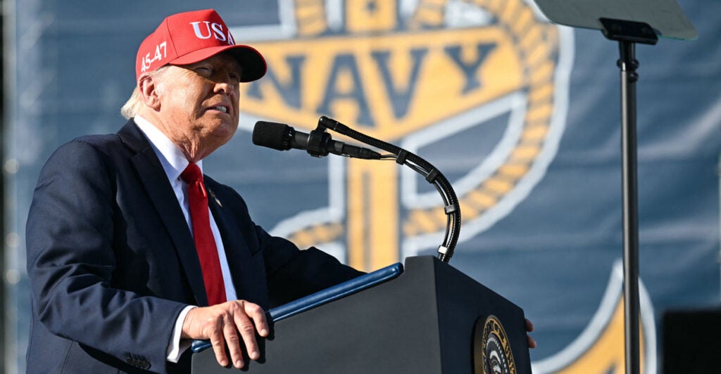 Donald Trump in suit and MAGA hat speaks, with Navy logo behind him.
