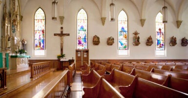 An empty church, looking across pews toward white wall with stain glass windows.