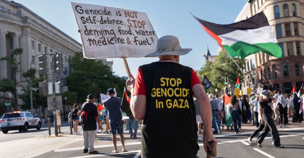 Man in black shirt with gold lettering reading "Stop Genocide in Gaza" and holding a sign walks down a Washington, DC street turning pro-Palestinian protest.