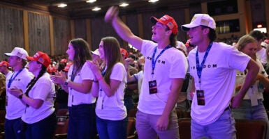 A group of young people in white shirts reading "Freedom" in black lettering cheering during Turning Point USA event.