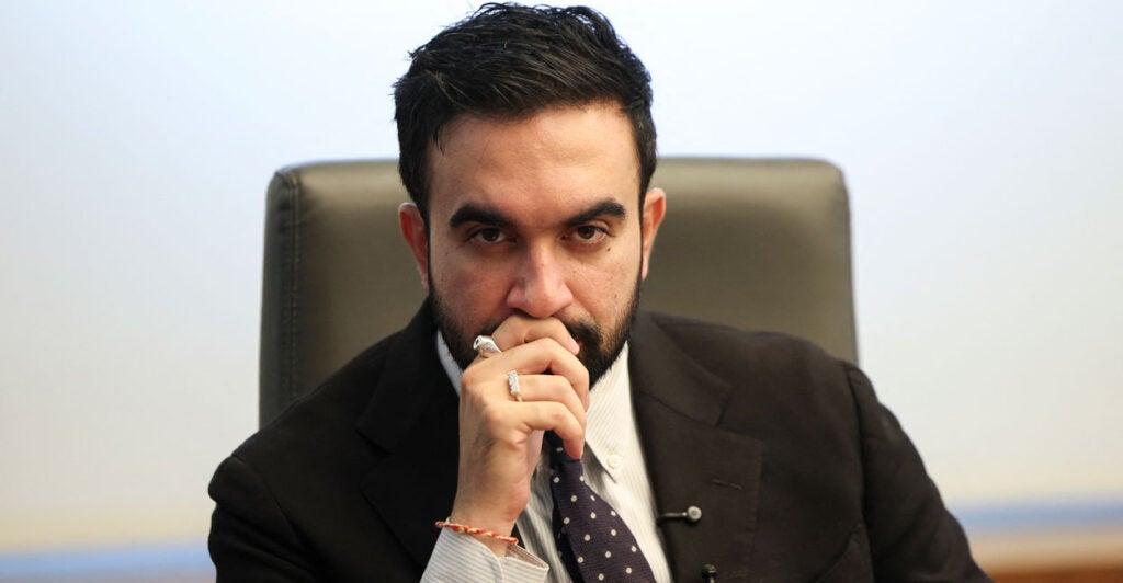 Zohran Mamdani staring into the camera lens, right hand at his mouth.