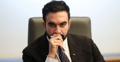 Zohran Mamdani staring into the camera lens, right hand at his mouth.