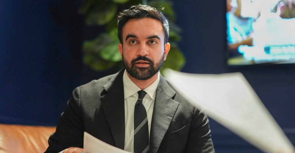 Zohran Mamdani in a dark suit and tie holds a paper in his hand.