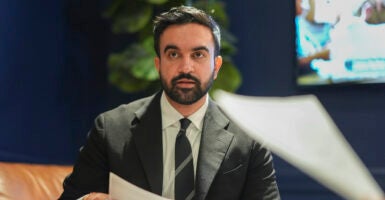 Zohran Mamdani in a dark suit and tie holds a paper in his hand.