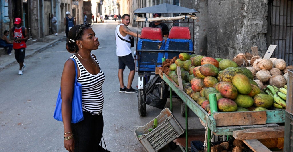 Cuban woman looks to by mangos at fruit stand, while man in background looks on.