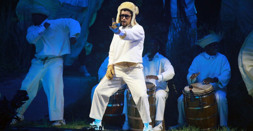 Bad Bunny, in a white outfit with fur hat, performs. Behind him, musicians with bongos.