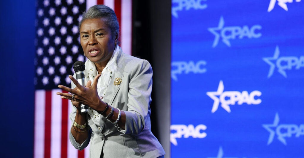 Winsome Earle-Sears in a gray suit stands in front of an American flag addressing an audience.