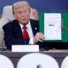 President Trump holds up the signed Gaza peace agreement, while sitting down.