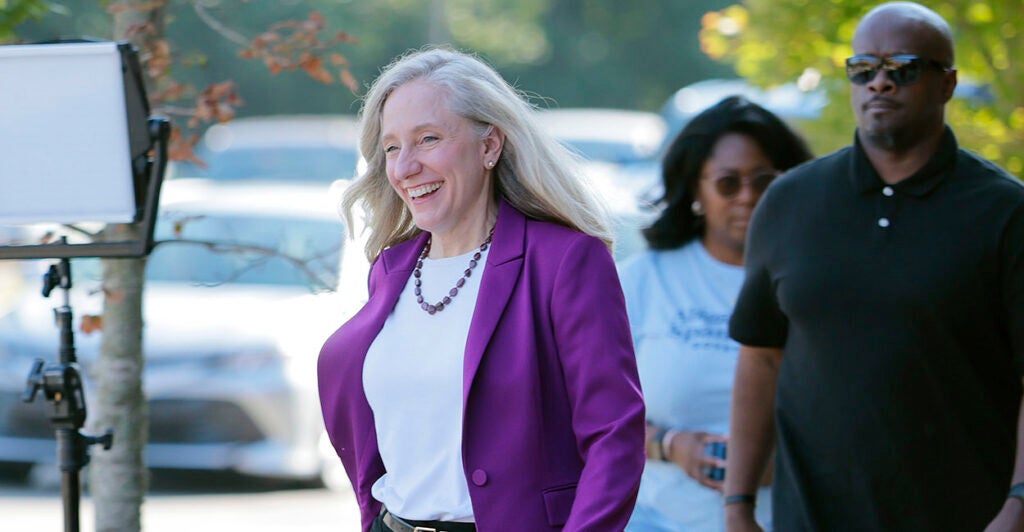 Democrat Abigail Spanberger smiles while wearing a white blouse with a blue sport coat in front of cars.
