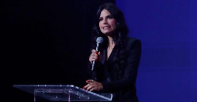 Turning Point USA podcast host Alex Clark on stage speaking into a handheld microphone in a black suit with a dark blue and black background