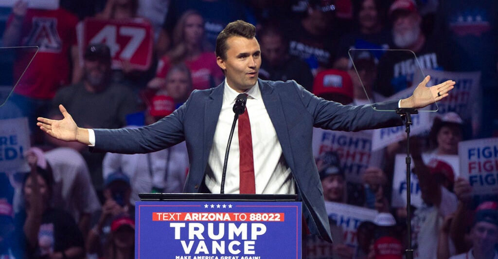 Turning Point USA founder Charlie Kirk in a blue suit with hands outstretched behind a Trump Vance sign.