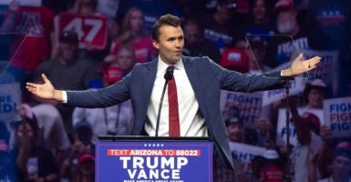 Turning Point USA founder Charlie Kirk in a blue suit with hands outstretched behind a Trump Vance sign.