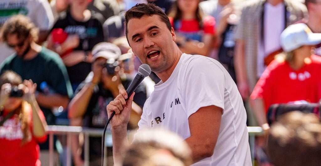 Charlie Kirk holds a microphone in a white shirt.